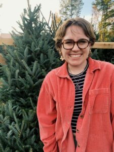 White woman with red glasses and jacket smiles while standing in front of a tree.
