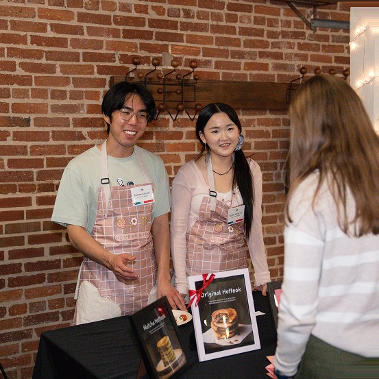 Two students in aprons presenting their hotteok stall to a visitor